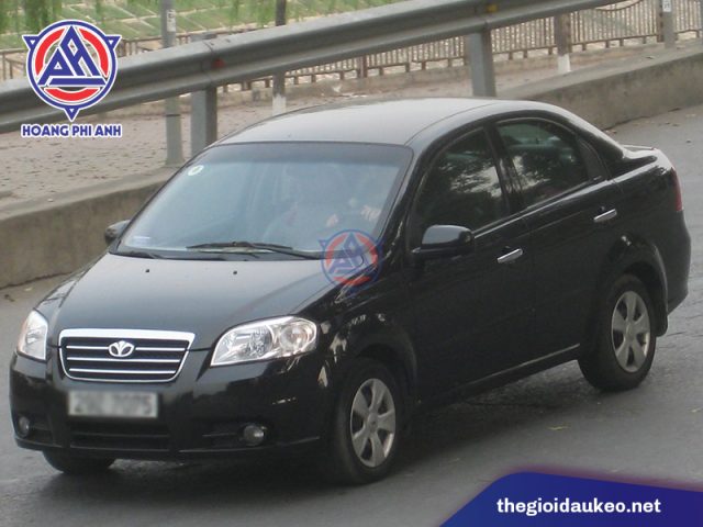 Daewoo Gentra màu trắng đang đỗ trên đường phố, phía trước có cửa hàng với tấm biển "Daewoo Gentra" và các ký tự khác